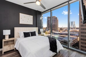 Une chambre moderne avec un grand lit, un ventilateur de plafond et une vue sur la ville grâce à de grandes fenêtres.