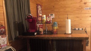 Et interiør af en træhytte med et barområde, baretter, en kaffemaskine og diverse madvarer på en hylde.