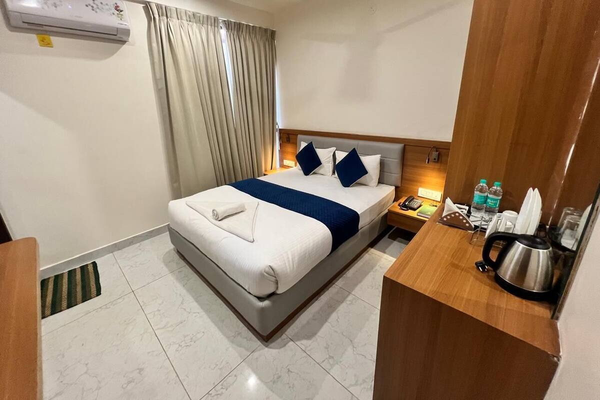 A hotel room with a bed, a bedside table with a kettle, a nightstand with a phone, and a window with curtains.