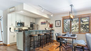 A modern kitchen with white cabinets, a central island with stools, a dining area with a table and chairs, and a large window with blinds. - Mountain Modern Ski In-Ski Out Condo With Hot Tub In Beaver Creek - 1BR/2BA (Beaver Creek)