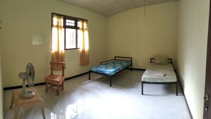 Ein Zimmer mit zwei Einzelbetten, einem Holzstuhl, einem Standventilator und einem Fenster mit Vorhängen.