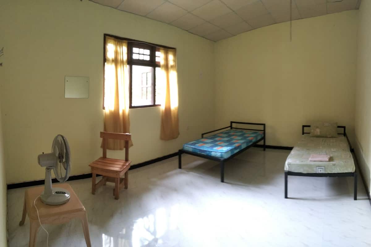 A room with two single beds, a wooden chair, a standing fan, and a window with curtains.