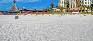 Una playa con una estructura de techo de paja, un edificio con techo rojo y un hotel de gran altura.
