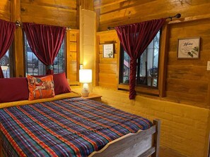 A wooden cabin room with a bed, red curtains, and a lamp.