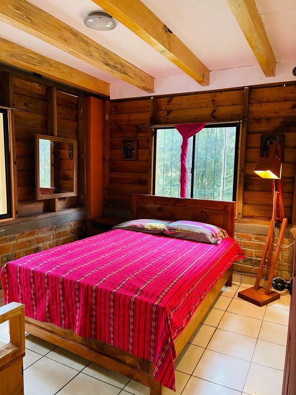 A wooden cabin bedroom with a bed, a window with curtains, a nightstand, and a lamp.