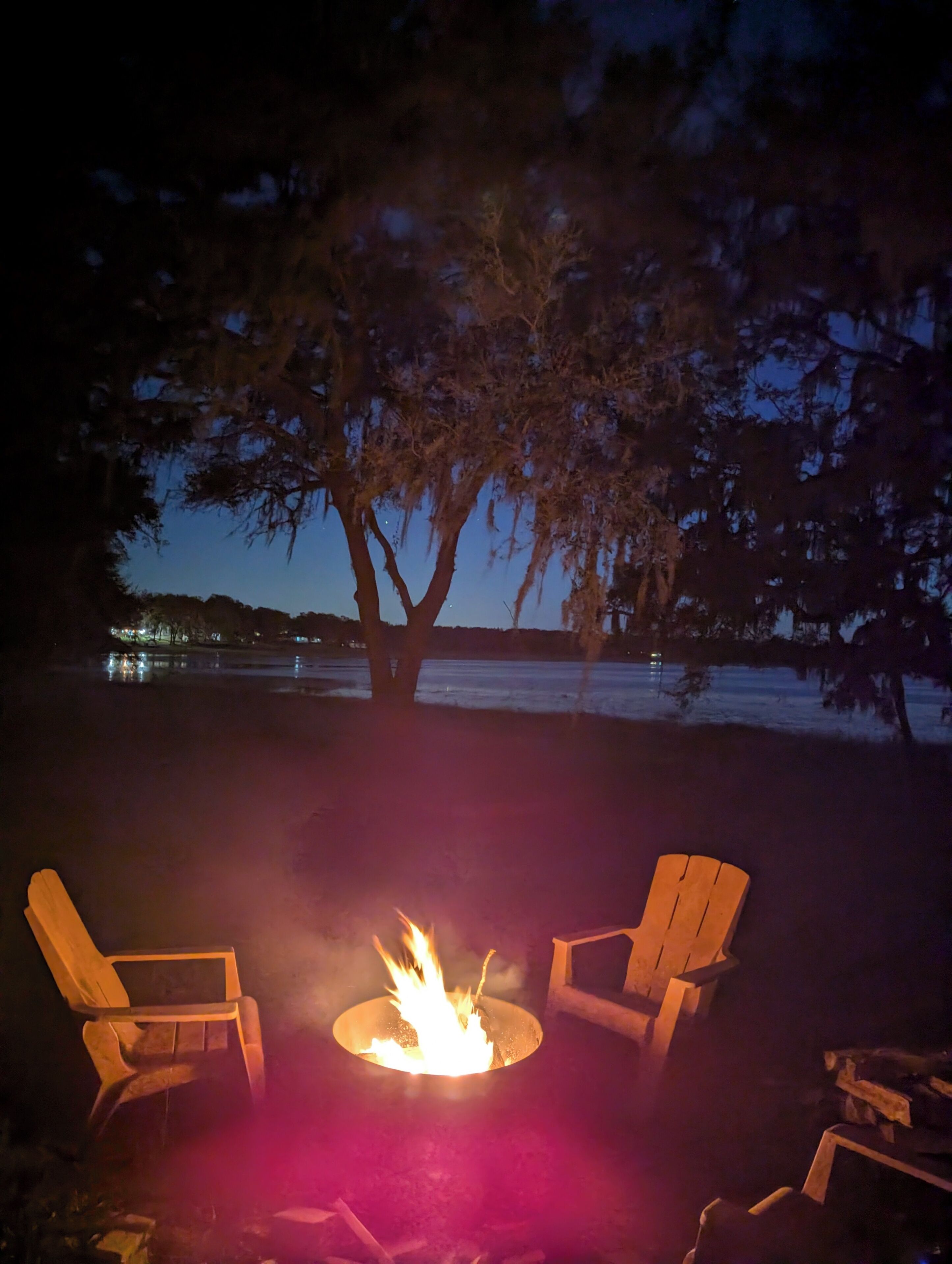 A nighttime outdoor seating area with a fire pit, two wooden chairs, and a large tree.