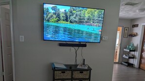 Un televisor montado en la pared muestra una vista panorámica de un lago. Debajo del televisor, hay un mueble de madera con cestas de mimbre.