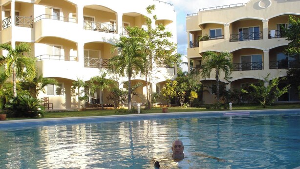 A swimming pool with a person in the water, surrounded by a building with balconies and palm trees.