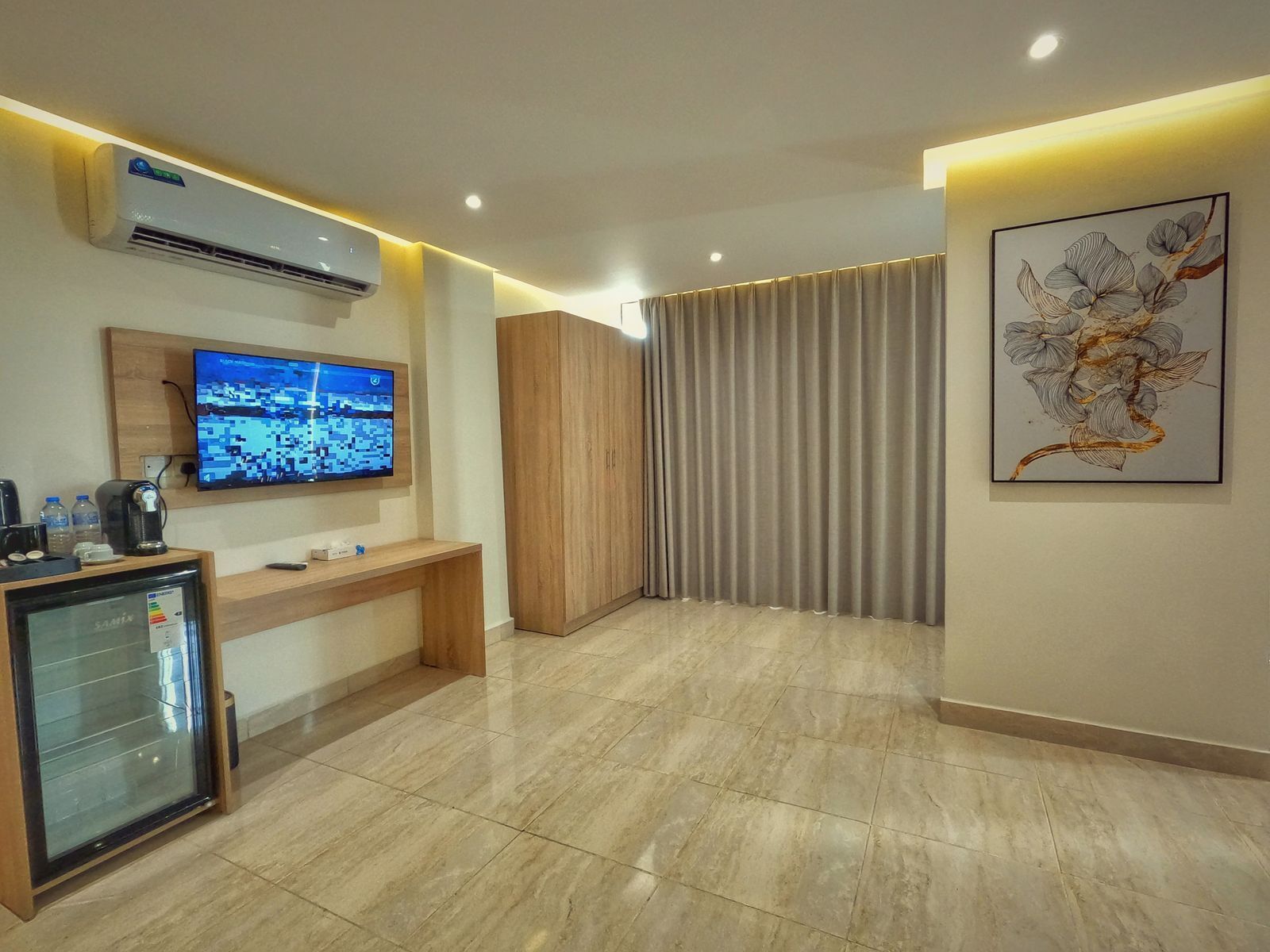 A hotel room with a large flat-screen TV, a wooden desk, a minibar, and a large window with curtains.