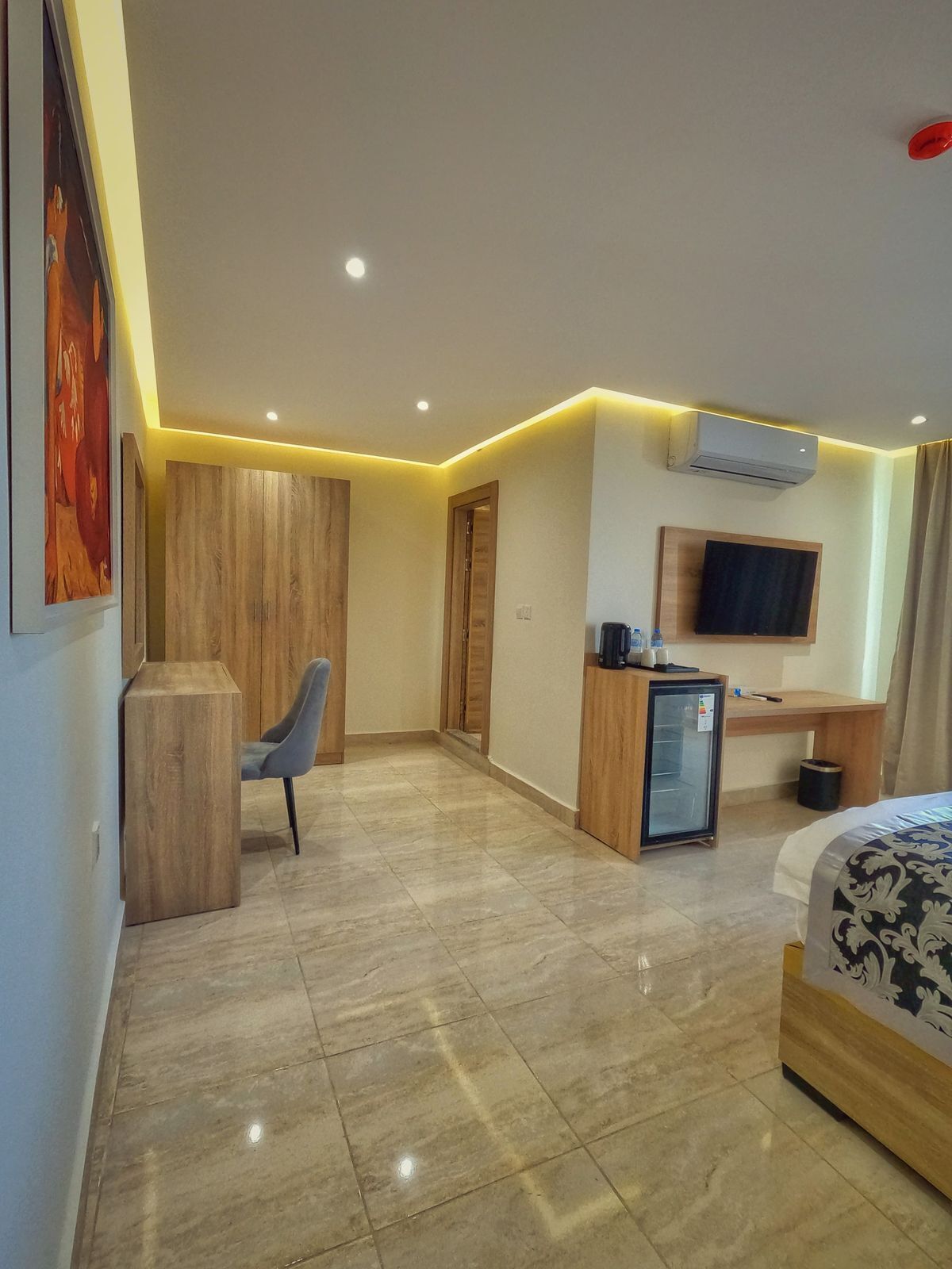 A hotel room with a bed, a desk with a computer, a chair, a small fridge, and an air conditioning unit.