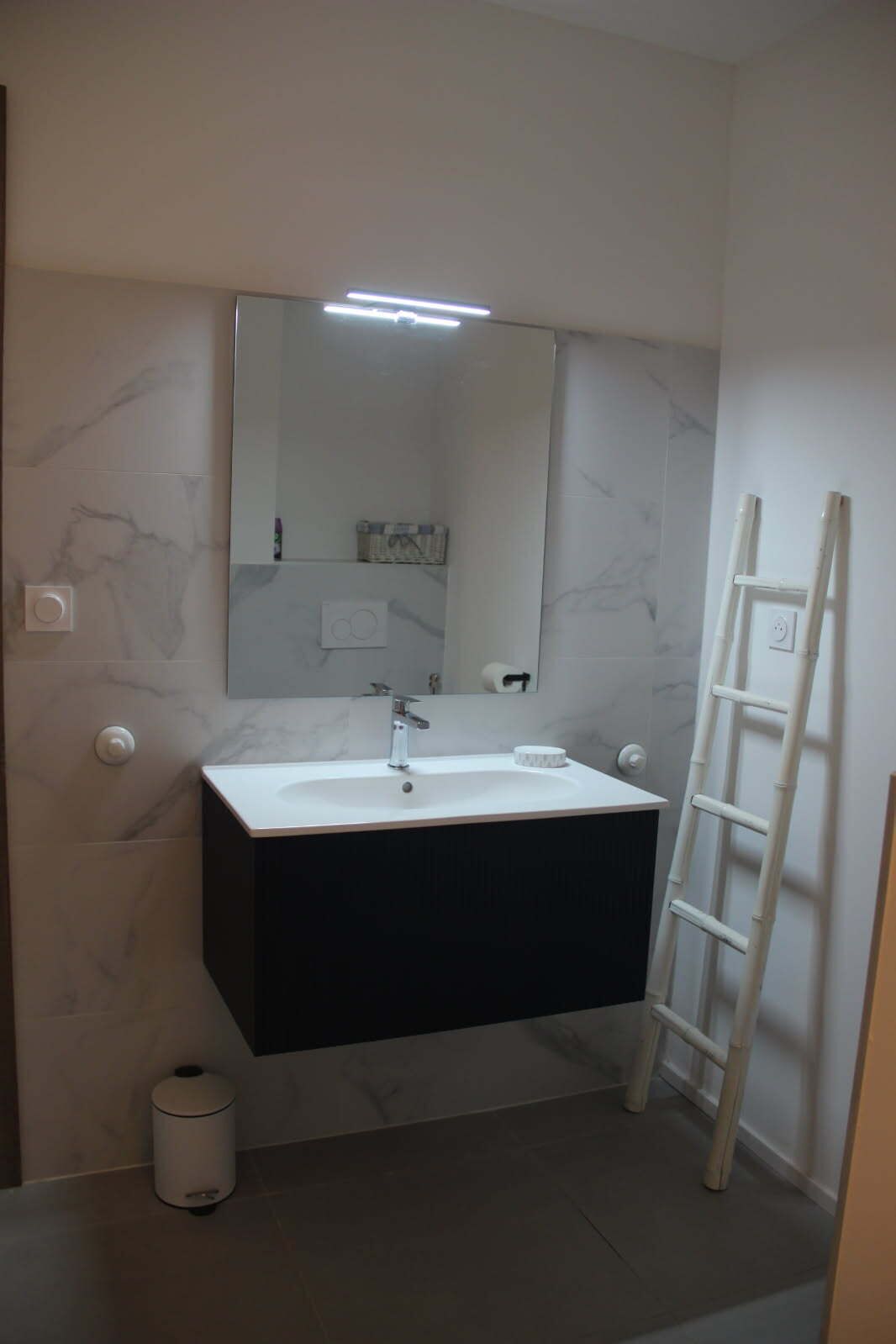 Un baño con un lavamanos blanco, un mueble negro, paredes de mármol, un espejo y una escalera blanca.