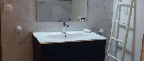 Un baño con un lavamanos blanco, un mueble negro, paredes de mármol, un espejo y una escalera blanca.