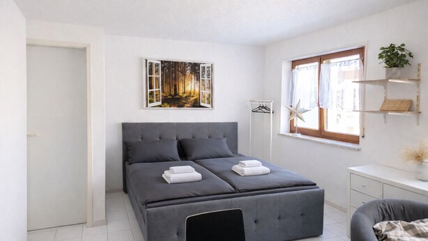 Un dormitorio moderno con una cama grande, una mesita de noche, una silla, una ventana con cortinas y un estante en la pared con una planta.