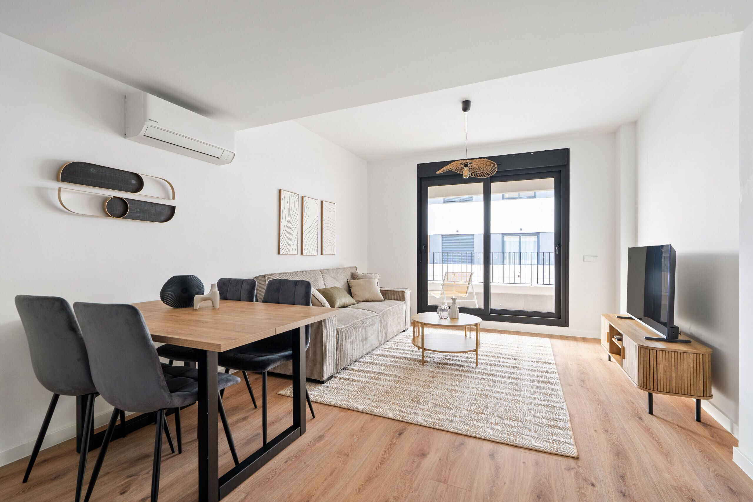 Un soggiorno moderno con un tavolo da pranzo in legno, sedie grigie, un divano, una TV e un'ampia finestra con vista.