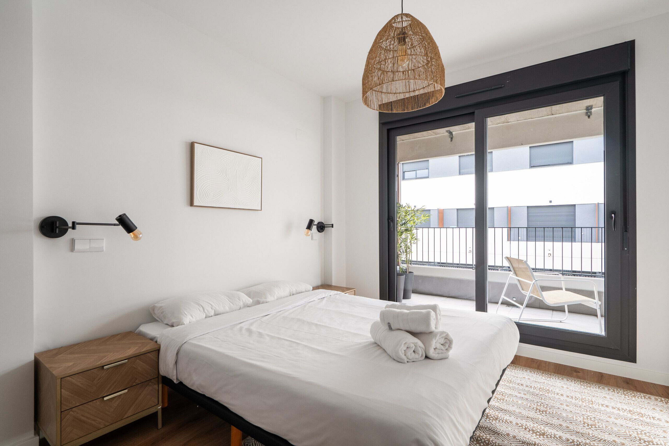 Una camera da letto con un letto, comodini, una lampada a sospensione, un quadro incorniciato e una porta scorrevole in vetro che dà su un balcone.