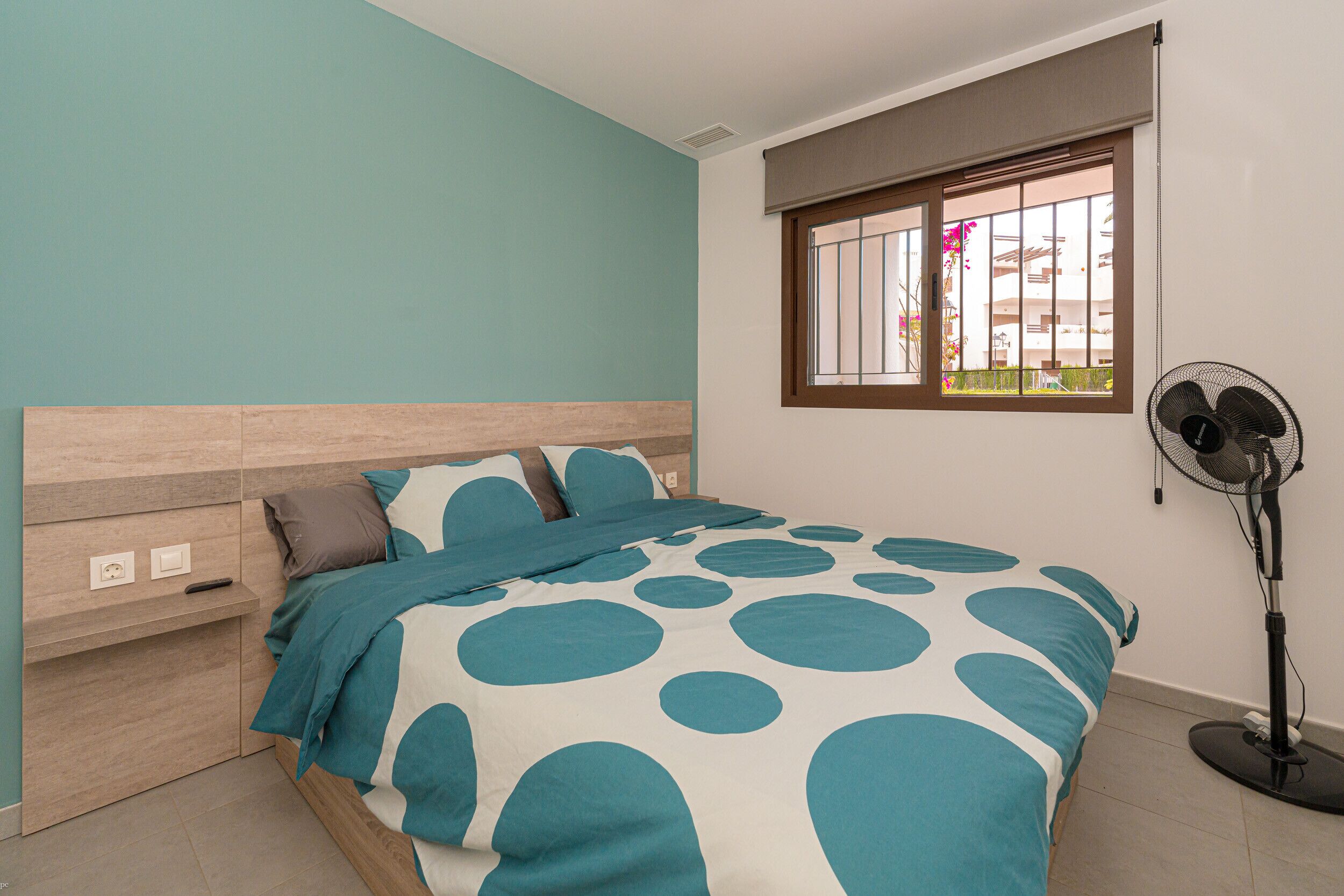 Ein Schlafzimmer mit einem Bett, einem Nachttisch, einem Fenster mit Blick und einem Standventilator.