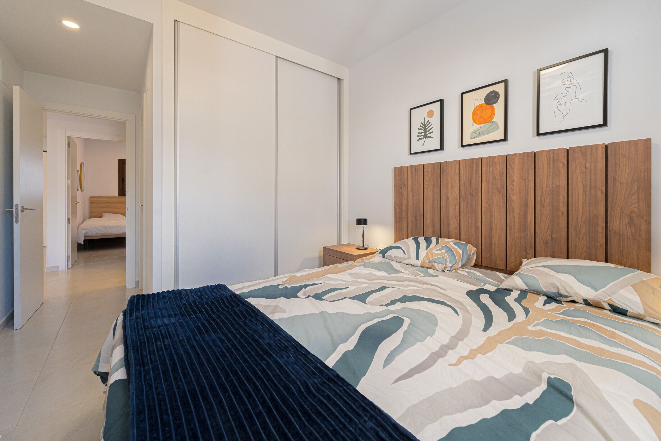 Un dormitorio con cama, cabecera de madera, mesita de noche y pared con tres cuadros enmarcados.