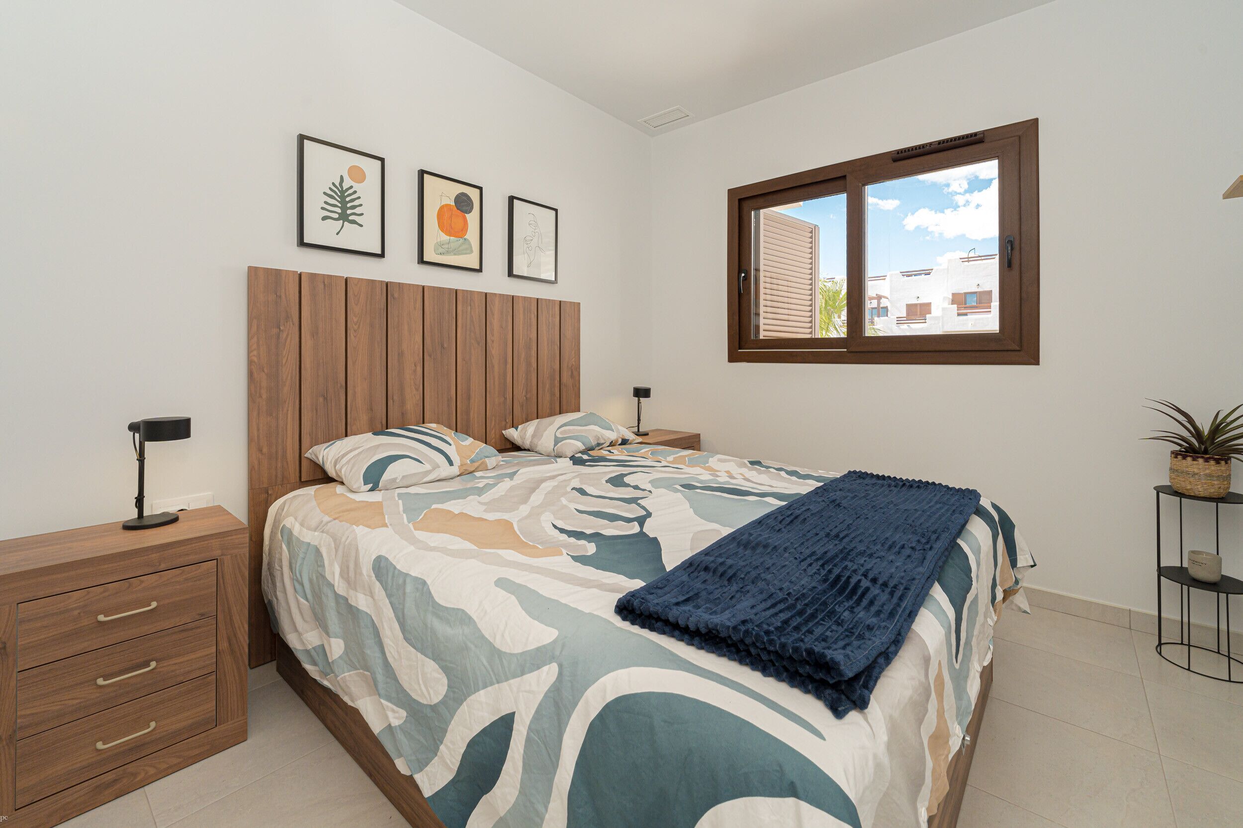 Une chambre à coucher avec une tête de lit en bois, un lit recouvert d’une couvre-lit à motifs, une fenêtre donnant sur des immeubles et une table de chevet avec une lampe.