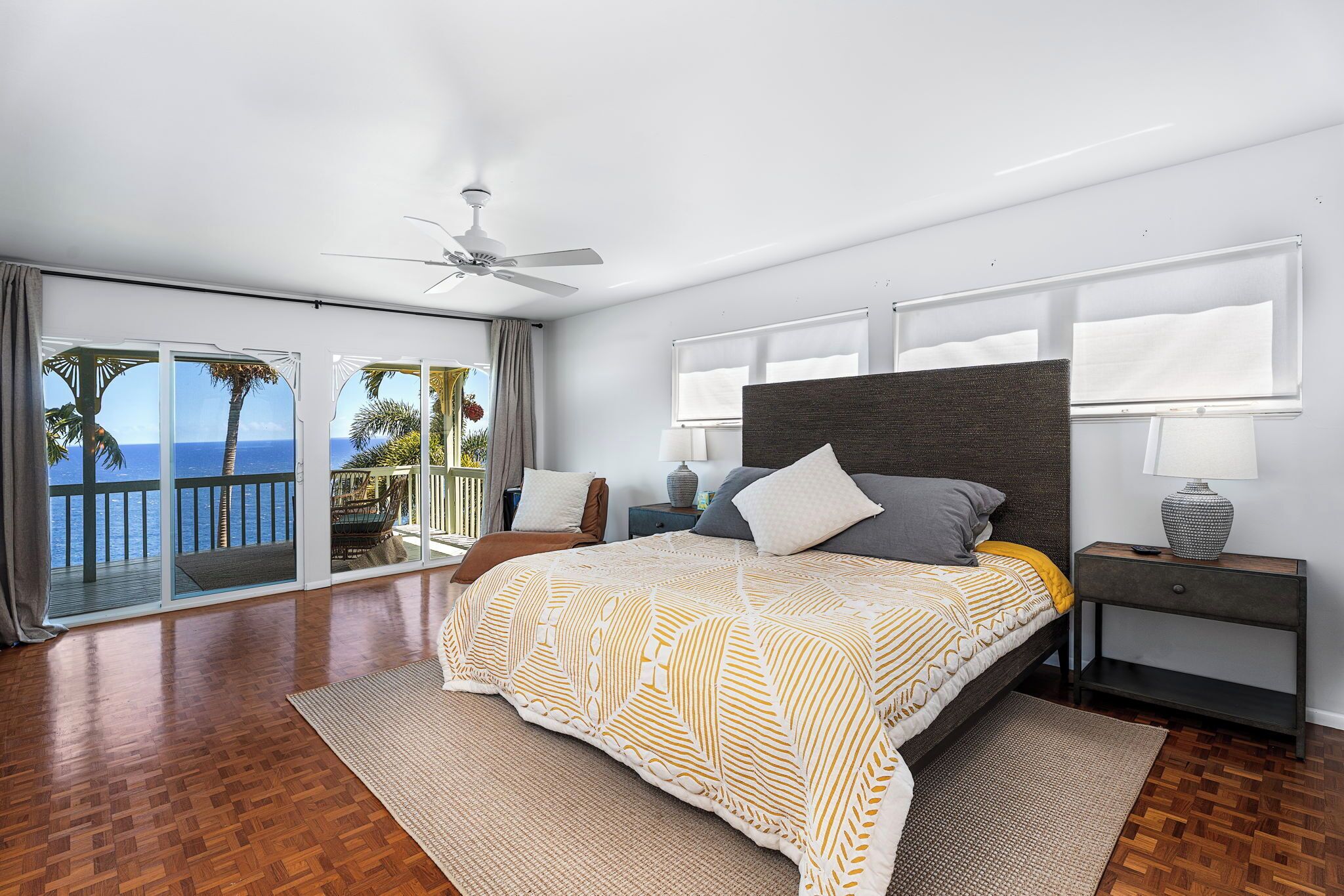 A modern bedroom with a large bed, a ceiling fan, and a view of the ocean through large sliding doors.
