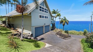 A light blue house with a garage, surrounded by a well-maintained lawn and palm trees, overlooking a clear blue ocean.