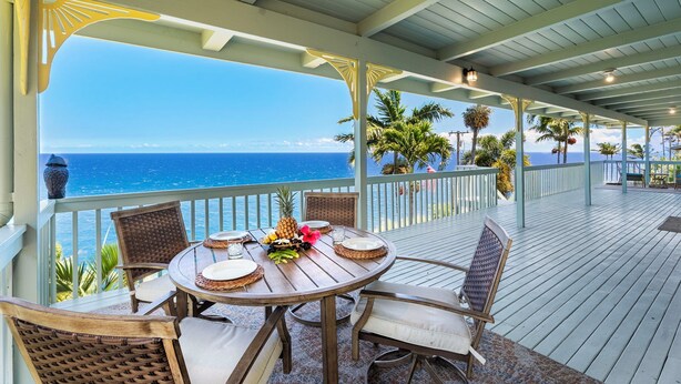 A covered outdoor dining area with a view of the ocean, featuring a round table with chairs and a tropical fruit centerpiece.