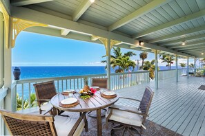A covered outdoor dining area with a view of the ocean, featuring a round table with chairs and a tropical fruit centerpiece. - Stunning Ocean-front Retreat Overlooking Laupahoehoe Point (LAUPAHOEHOE)