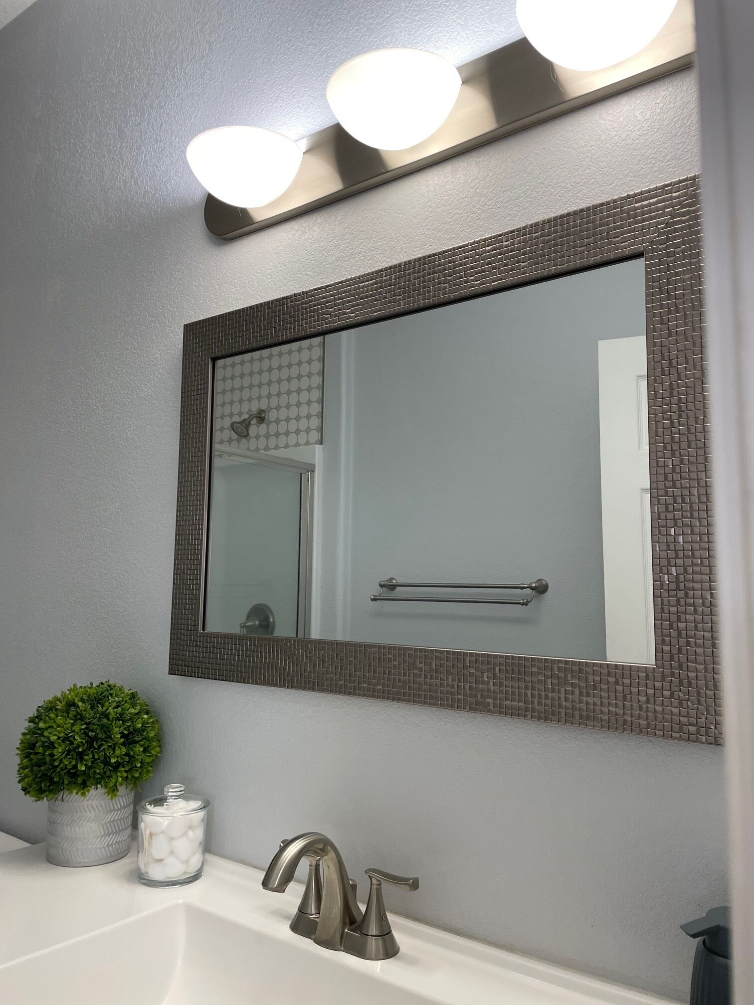 Un baño con un espejo grande, tres luces empotradas, un lavabo con grifo moderno, una pequeña planta en maceta y un frasco de vidrio.