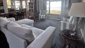 A spacious living room with a high ceiling, large windows, and a view of the outdoors. There is a fireplace, a dining area, and a seating area with a sofa and chairs.