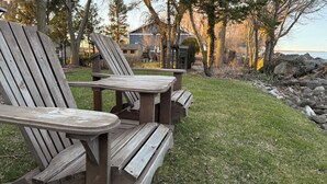Two Adirondack chairs and a picnic table in a grassy outdoor area with a view of a house and trees.
