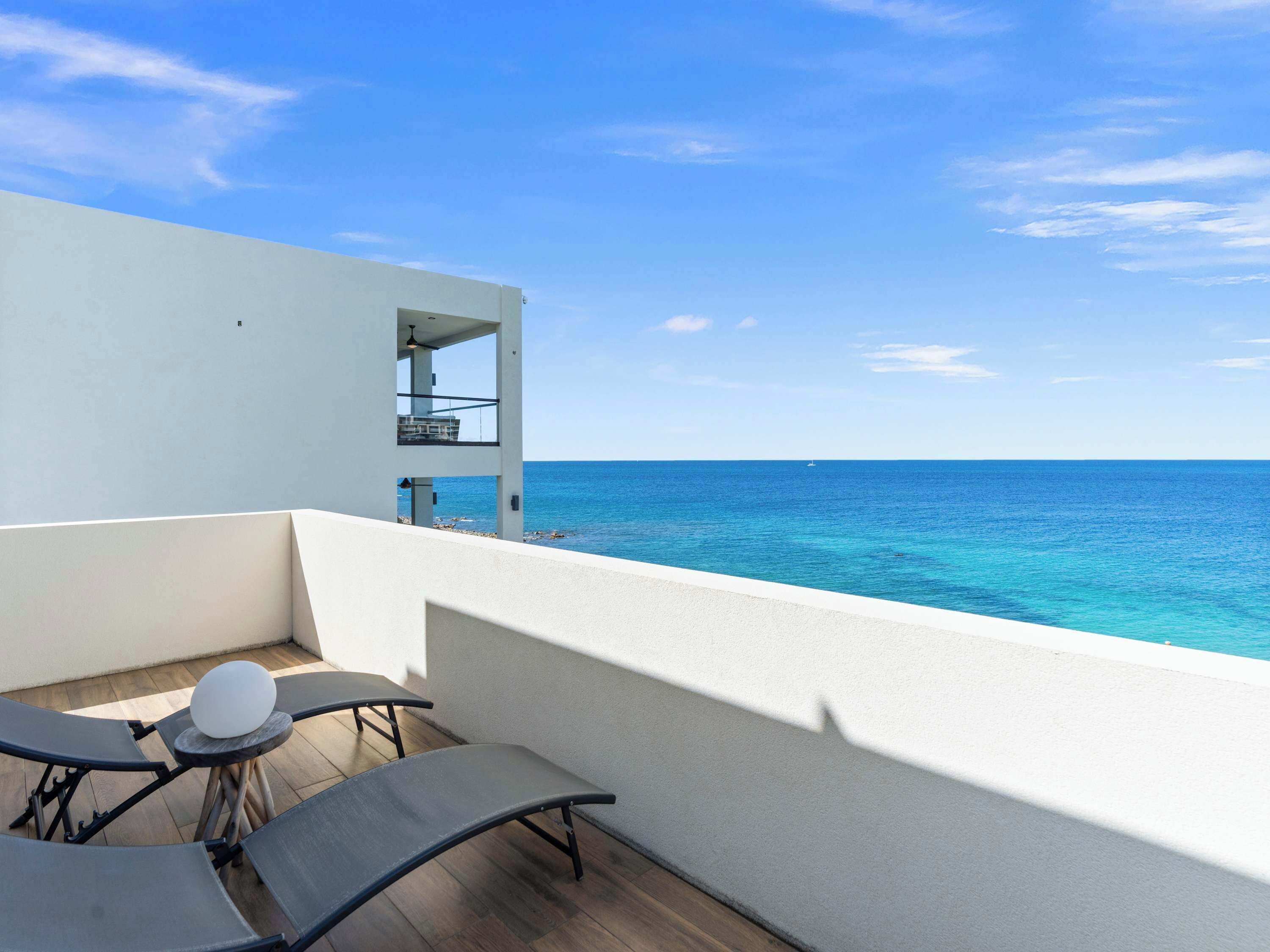 A balcony with a view of the ocean, a white building, and two lounge chairs.