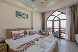 Ein Hotelzimmer mit einem großen Bett, einem Schreibtisch, einem Sessel, einem Balkon mit Blick und einem Bild an der Wand.