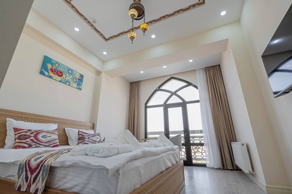 A hotel room with a bed, a ceiling light fixture, a window with curtains, and a radiator.
