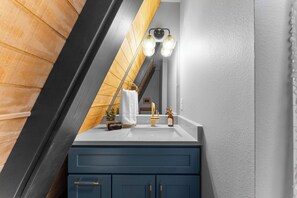 Ein Badezimmer mit weißer Ablagefläche, einem goldenen Wasserhahn und einem Spiegel mit drei Lampen.