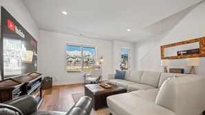 A modern living room with a fireplace, a large window offering a mountain view, and a wall-mounted TV displaying streaming services. - Terrapin Basin R5939 | Ski Escape | Hot Tub & Desk (Mountain Green)
