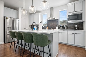 A modern kitchen with a central island, bar stools, stainless steel appliances, and a view of the outdoors. - Elliott’s Escape R5977 | Snowbasin Skiing |Hot Tub (Mountain Green)