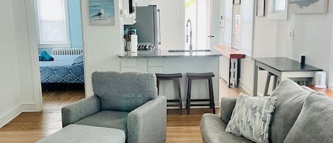 A modern living room with grey sofas, a wooden floor, and a kitchen area with white cabinets and a ceiling fan.