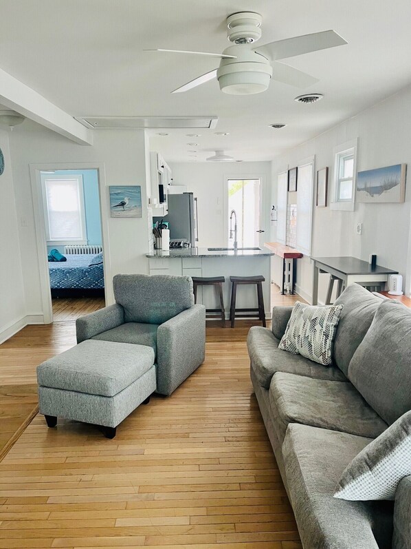A modern living room with grey sofas, a wooden floor, and a kitchen area with white cabinets and a ceiling fan.