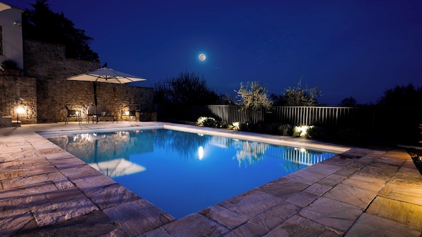 A swimming pool with stone tiles, a patio area with chairs and an umbrella, and a stone wall in the background.