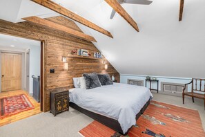 A bedroom with a sloped ceiling, wooden beams, a bed with white bedding, a red and orange rug, and a small table with decorative items. - Captain King's Farm by Kristin and Sakonnet Farm & Stays (Tiverton)