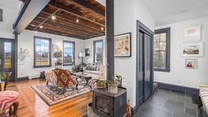 A spacious living room with a large black wood-burning stove, wooden floors, and a high ceiling with exposed beams. There are multiple windows providing natural light, and the room is decorated with various plants and artwork.