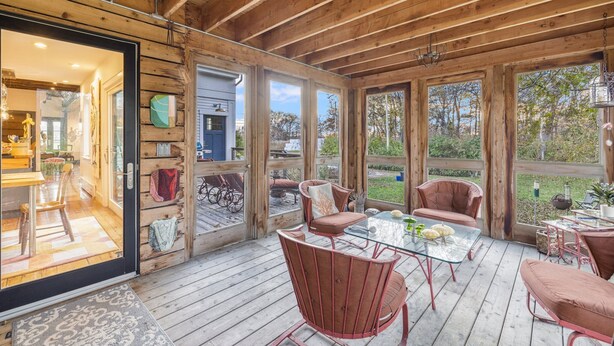 A wooden deck with glass doors, furnished with red chairs and a glass table, overlooking a garden.