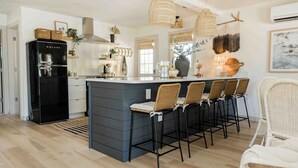 A modern kitchen with a central island, bar stools, a black refrigerator, wicker chairs, and pendant lights.