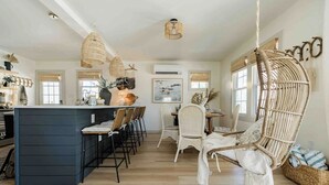 A modern kitchen with a central island, bar stools, wicker chairs, and pendant lights.