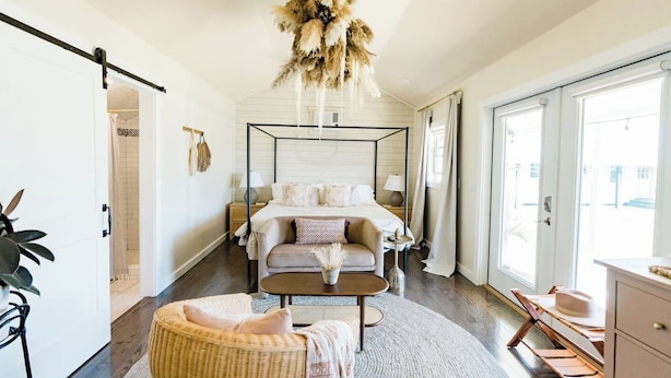 A bedroom with a four-poster bed, a round rug, a wicker chair, and a view of the outside through a large window.