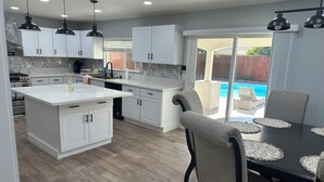 A modern kitchen with white cabinets, a large island, and a dining area with a pool view. - Spacious cozy Oasis retreat with private pool  Backyard (Modesto)