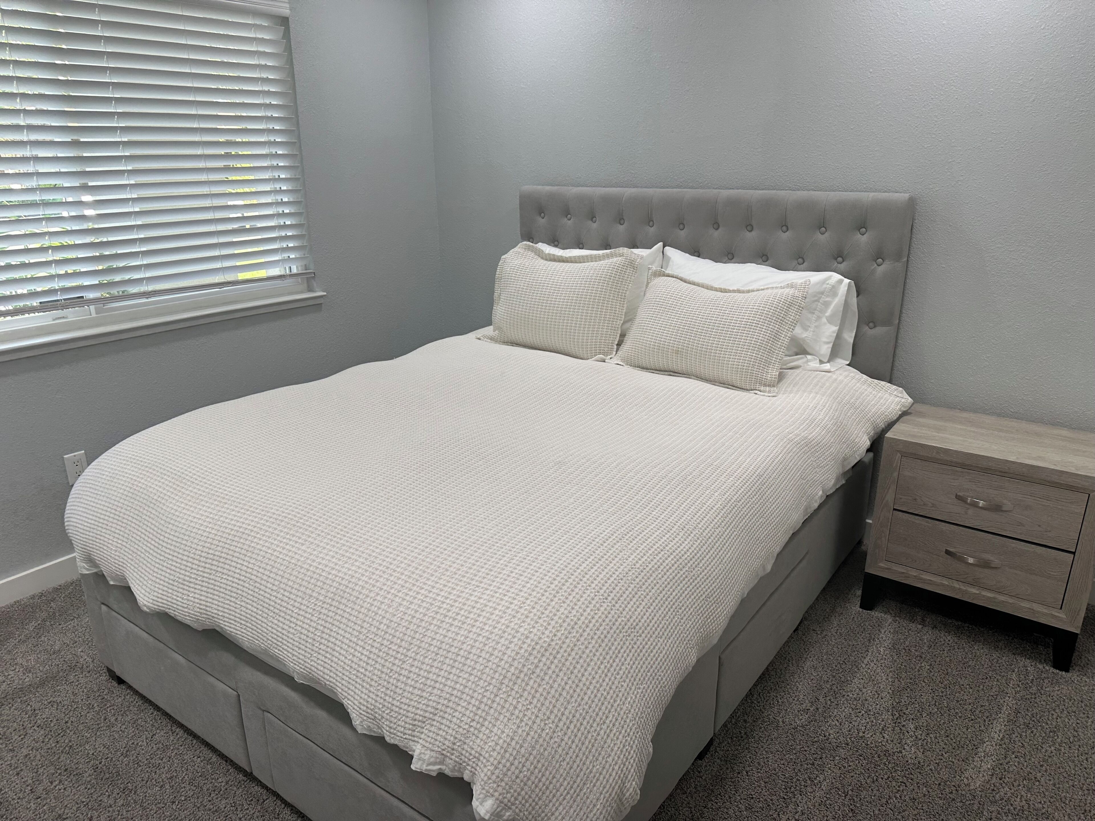 Un dormitorio con cabecera capitonada, una cama grande con colcha blanca texturizada, dos almohadas, una mesita de noche con cajón y una ventana con persianas.
