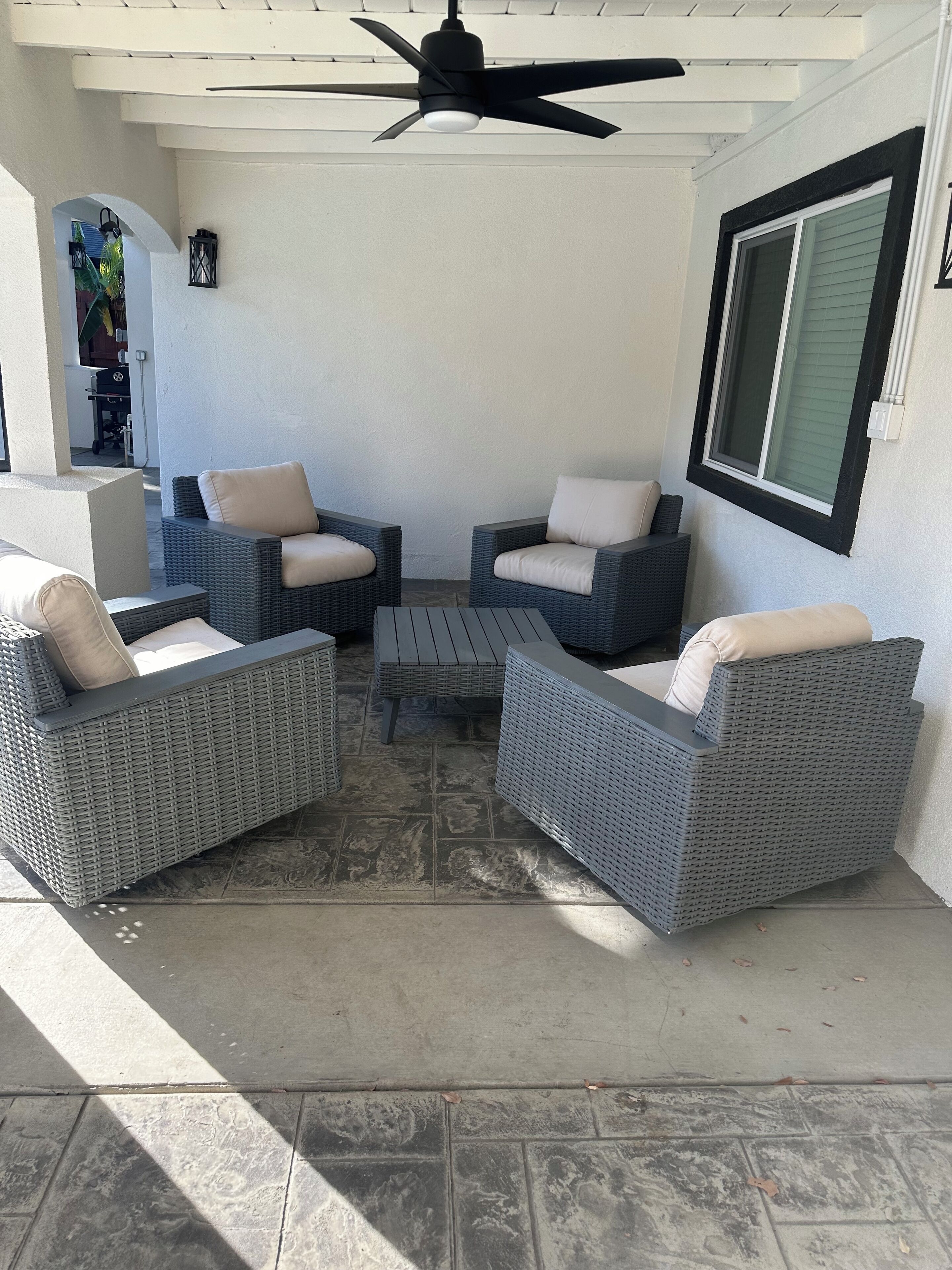 Un espace patio aménagé avec des meubles en osier, un ventilateur de plafond et une fenêtre équipée de stores.