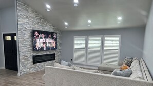 A living room with a stone wall, a large flat-screen TV, a grey sofa, and a window with blinds. - Spacious cozy Oasis retreat with private pool  Backyard (Modesto)