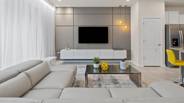 Ein modernes Wohnzimmer mit Sofa, Couchtisch, Fernseher und Küchenbereich mit Kühlschrank und gelbem Sessel.