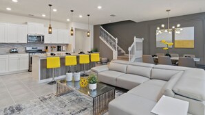 Eine moderne Küche mit weißen Schränken, einer zentralen Kochinsel mit gelben Barhocker, einem Essbereich mit grauem Sofa und einer Treppe zu einer zweiten Etage.